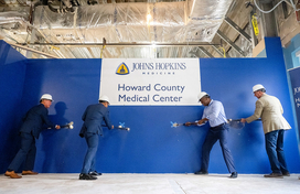 A photo of County Executive Calvin Ball and others swinging sledge hammers into a big blue wall to ceremoniously break the wall on the new observation unit at Johns Hopkins Howard County Medical Center.