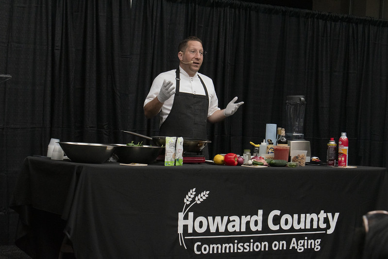 Chef Egg behind cooking demo table with tablecloth saying Howard County Commission on Aging