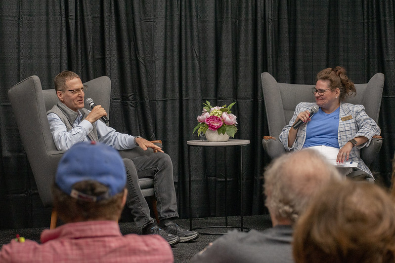 Male and female sitting in comfy chairs talking and holding microphones.