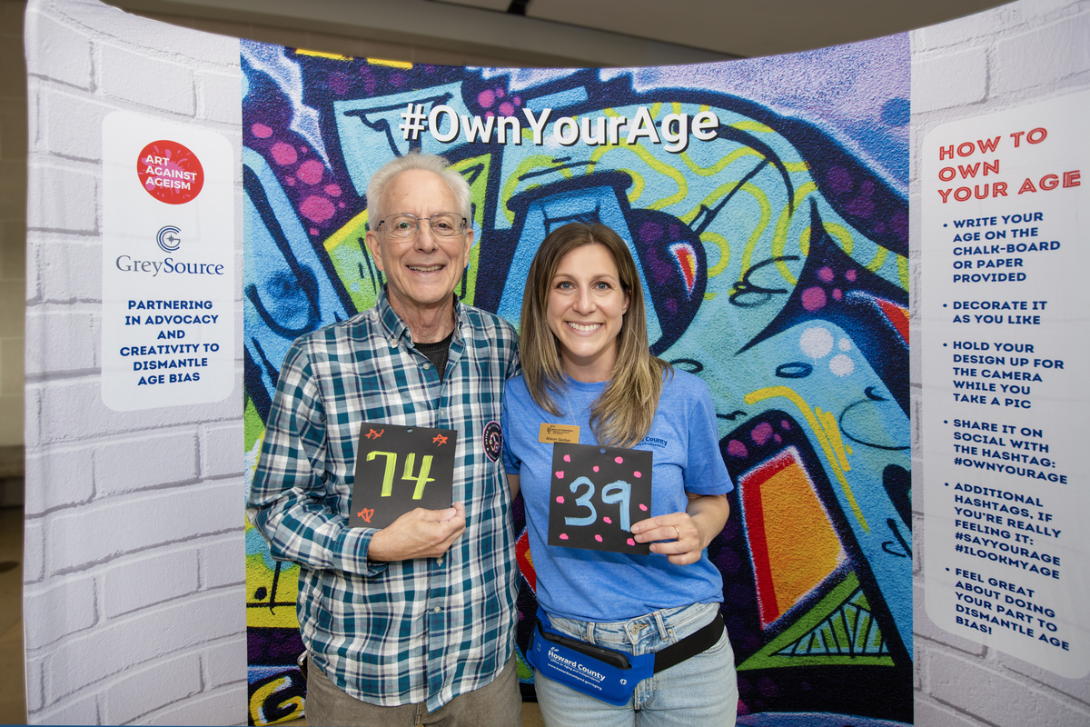Older white male and middle aged female posing for photo together at Own Your Age Photobooth. The male is holding a sign saying 74 and female is holding a sign saying 39.
