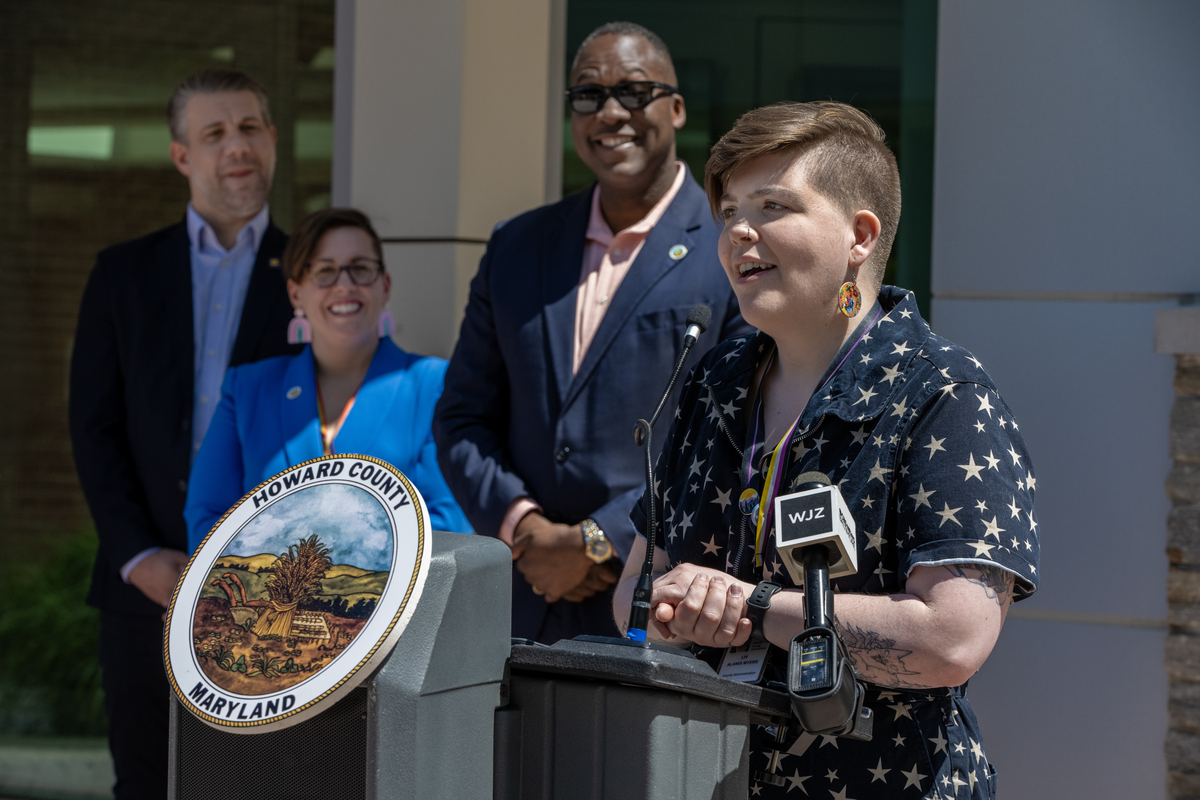 A person with a short undercut speaks into a microphone at a podium. 