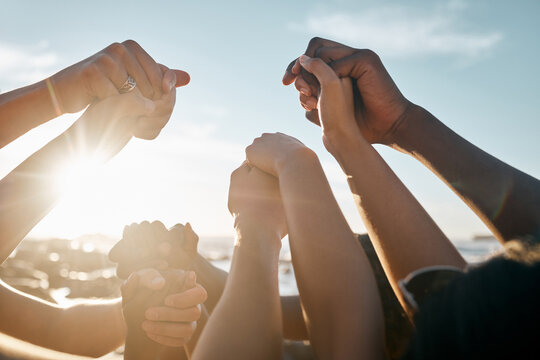 Multiple people raise their clasped hands together in a circle against a bright sky, symbolizing unity, support, and community.
