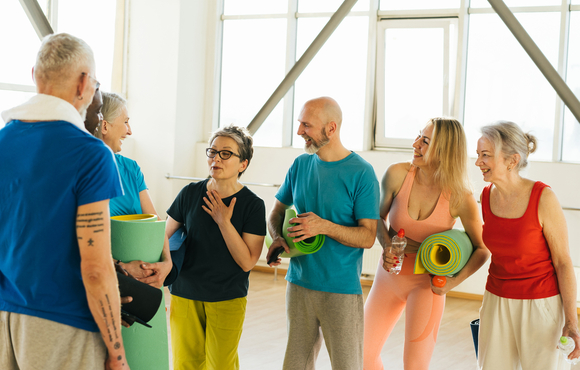 Group of 6 older adults in exercise outfits and some holding yoga mats. There are 2 men and 4 women. They look happy and chatting with each other.