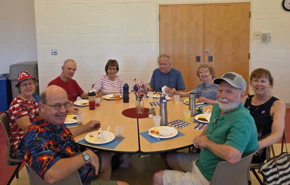 Group of eight older adults sitting around a round table eating lunch together and smiling.