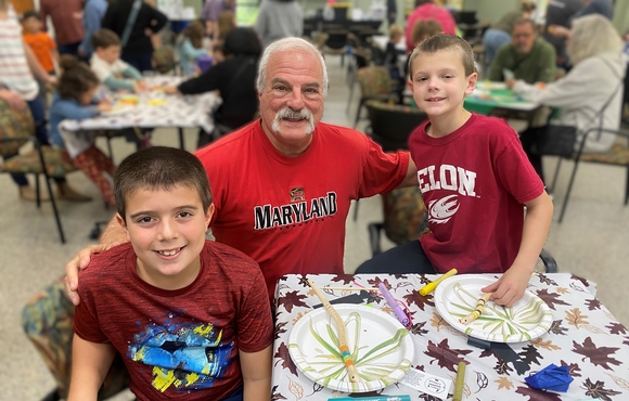 Older adult white male with two kids elementary school age, all wearing red t-shirts posing together smiling at an arts & crafts table.