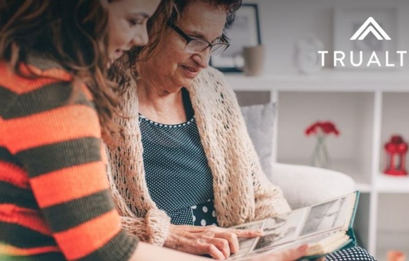 Two women, one older and one younger, sitting on a couch together looking over a handout. Trualta logo in the upper right hand corner of the photo.