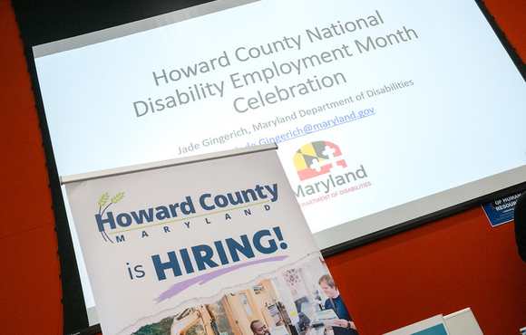 A photo of a project screen that reads Howard County National Disability Employment Month Celebration with a pop up sign in front of it that reads Howard County is hiring.