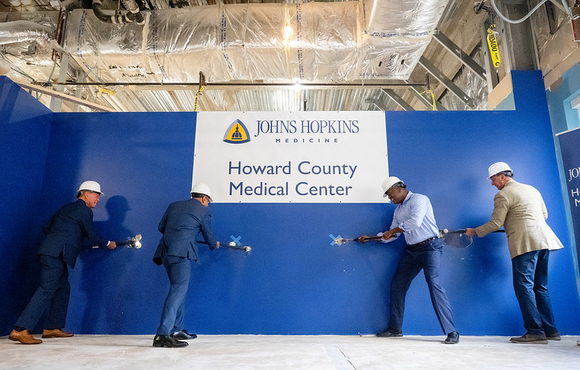 A photo of County Executive Calvin Ball and others swinging sledge hammers into a big blue wall to ceremoniously break the wall on the new observation unit at Johns Hopkins Howard County Medical Center.