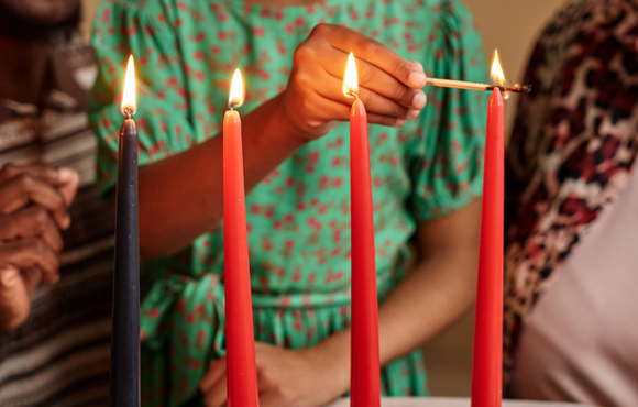 Two adults watch as a child lights a red candle on a Kwanzaa kinara