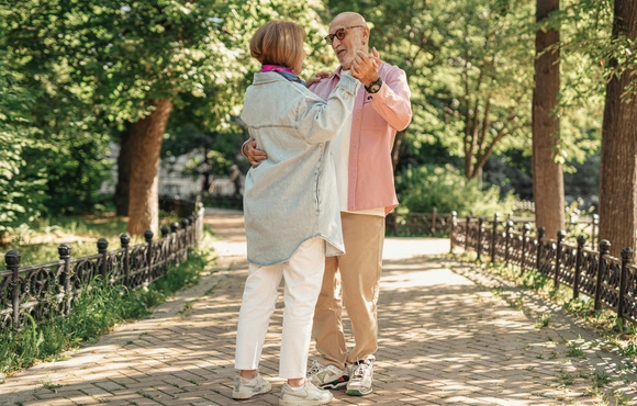 Older adult couple dancing on a sidewalk in a park on a nice day.