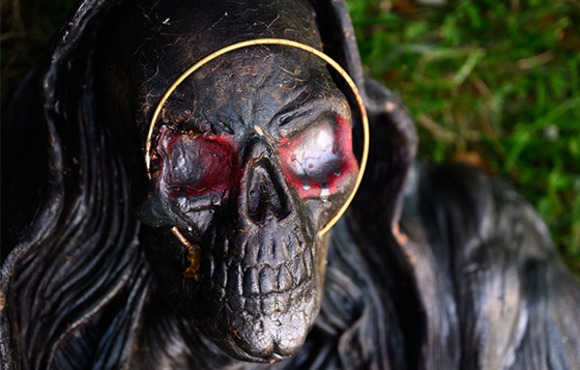 A close-up of a dark, weathered skull figure wearing a black hood, featuring red-painted eye sockets and a thin gold ring encircling its face against a green grass background.