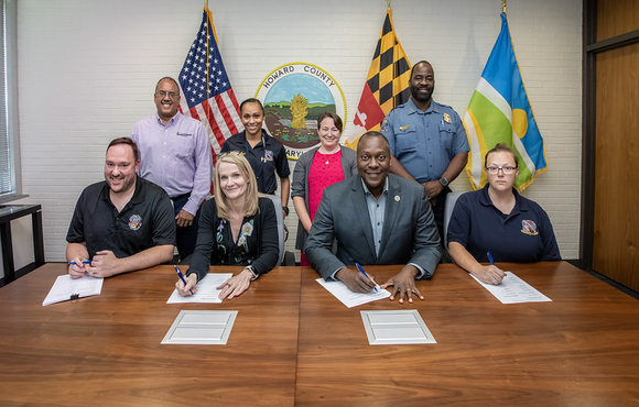 County Executive Calvin Ball seated at a table with the County's Chief Administrative Officer Brandee Ganz and members of Howard County Government and Howard County Public Dispatcher’s Association, with other officials standing behind, signing a memorandum of agreement between the County and the group.