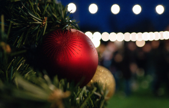 green christmas tree branches in the foreground adorned with a gold ball and a red ball ornament and white string lights glowing the background