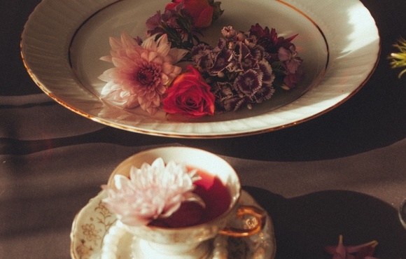 a small white teacup filled with red and white flowers on a white saucer sitting surrounded by a string of pearls near a bowl filled with white, red, and brown flowers all on a brown wooden table adorned with flower petals