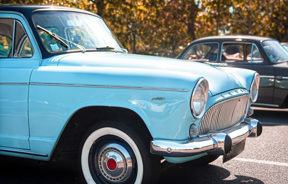 an older model turquoise car is parked by a black car a couple stalls away with golden-hued trees in the background