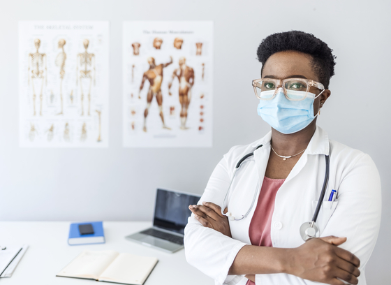 Nurse standing in exam room wearing a mask. Anatomy posters and laptop in the background