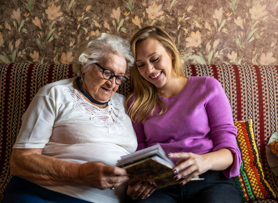 Older woman with white hair and glasses looking at a photo album on a couch with a younger woman with blonde hair.