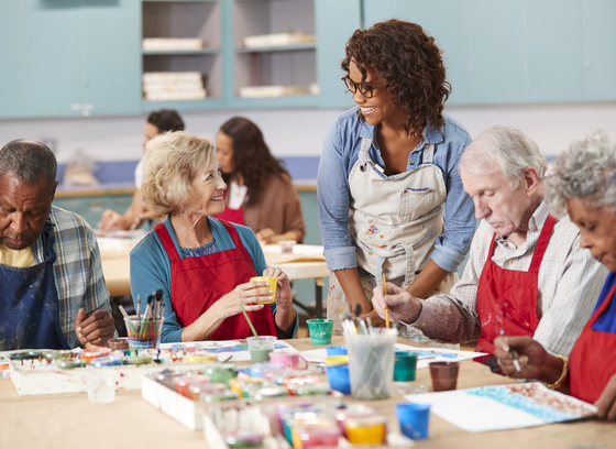 Group of 4 older adults in a painting class with a female instructor talking to them at their table,