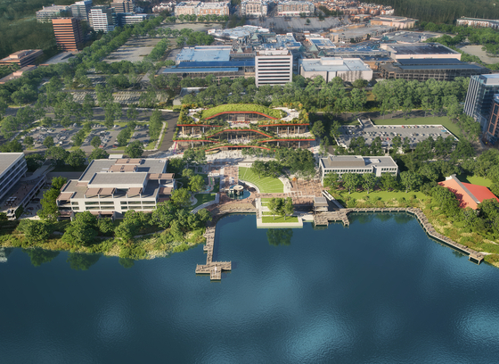 Aerial view of a modern lakeside urban area with office buildings, landscaped green spaces, and wooden boardwalks along the water. A large, architecturally distinct building with terraced green roofs sits at the center, surrounded by trees, parking lots, and additional structures. The calm blue lake in the foreground reflects the greenery, while a mix of commercial and residential buildings extends into the background.