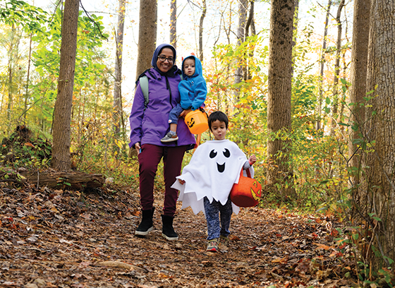 A child wearing a ghost costume and holding a trick-or-treat basket walks along Robinson Nature Center trails followed by his smiling mother and sibling