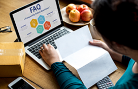 A person looking at a paper while an FAQ page is open on their laptop