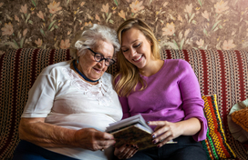 Older woman with white hair and glasses looking at a photo album on a couch with a younger woman with blonde hair.