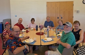 Group of eight older adults sitting around a round table eating lunch together and smiling.