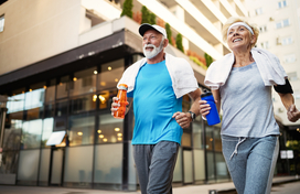 Older adult man and woman walking together in workout clothes holding water bottles and towels around neck.