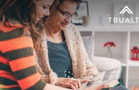 Two women, one older and one younger, sitting on a couch together looking over a handout. Trualta logo in the upper right hand corner of the photo.