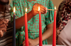 Two adults watch as a child lights a red candle on a Kwanzaa kinara