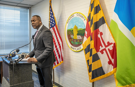 A photo of County Executive Calvin Ball standing at a podium inside his conference room in the George Howard Building, with the County seal and United States, Maryland, and Howard County flags behind him during his briefing to the media about Howard County Government revoking a building permit for a proposed detention center in Elkridge.