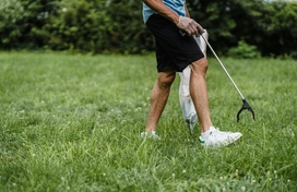 Man picking up trash on grass with a grabber; only abdomen to feet showing.