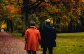 man and woman walking surrounded by leaves on the ground and trees changing to autumnal colors