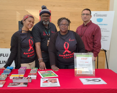 A group of four adults standing behind a table at a public awareness booth. Three of them wear black shirts with red ribbons and the words ‘Awareness Rocks,’ suggesting HIV/AIDS awareness. The table is covered with brochures, pamphlets, and a display sign that reads ‘STOP HIV.’ One person stands slightly apart in a red shirt. The group appears to be at an indoor event or community outreach setting