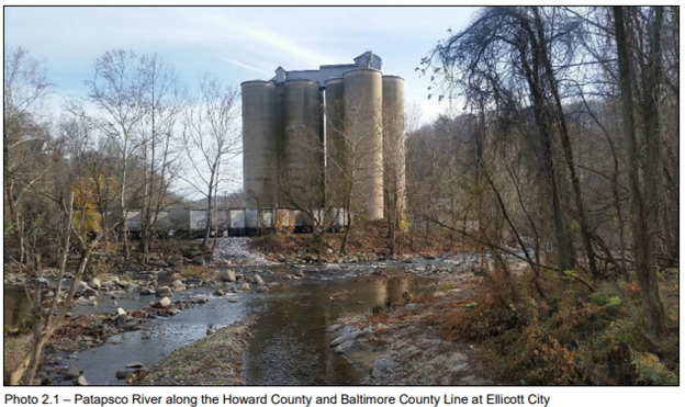 Four large concrete silos stand beside the Patapsco River, surrounded by leafless trees, with rocky riverbanks in the foreground.