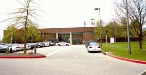 Street-view style image of a large brick building with a central entrance and parking area in front. There are a few parked cars, leafless trees, grassy areas, and black lamp posts lining the driveway. The sky appears overcast.