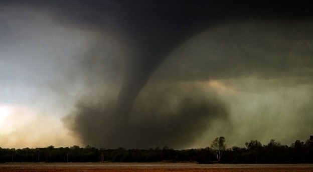 A large tornado extends from a dark storm cloud to the ground across a flat landscape. Dust and debris swirl around the base of the funnel, while the sky appears gray and ominous, with trees lining the horizon in the background.