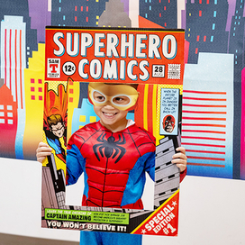 Kid in spiderman costume holding posing for a photo while holding a cardboard frame saying "Superhero Comics"