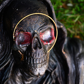 A close-up of a dark, weathered skull figure wearing a black hood, featuring red-painted eye sockets and a thin gold ring encircling its face against a green grass background.